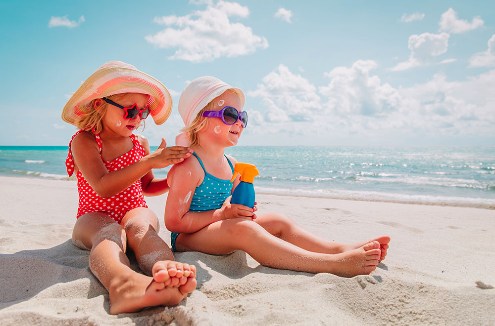 Dos chicas en la playa en traje de baño, gafas de sol y sombreros, aplicándose protector solar.