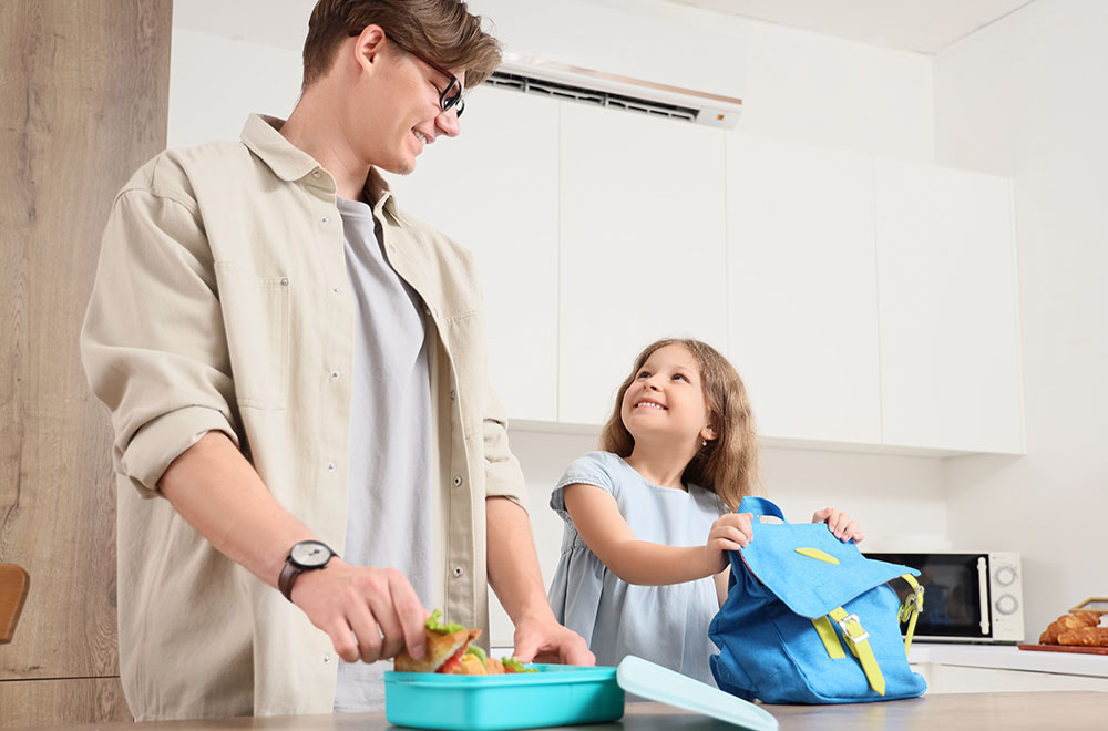 El padre está preparando el almuerzo para su hija, quien sonríe mientras sostiene la mochila.