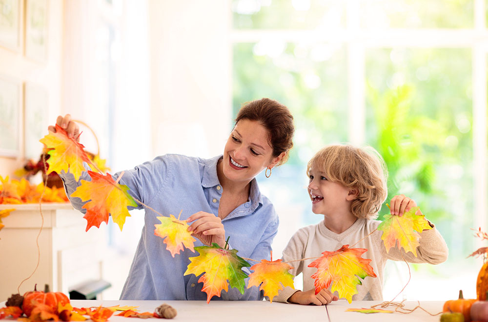 Mamá e hijo crean una guirnalda de hojas de colores, sonriéndose en una habitación clara y soleada.