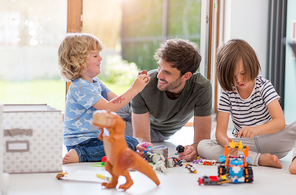Un padre juega con sus hijos en el suelo, organizando juntos los juguetes en una habitación soleada.