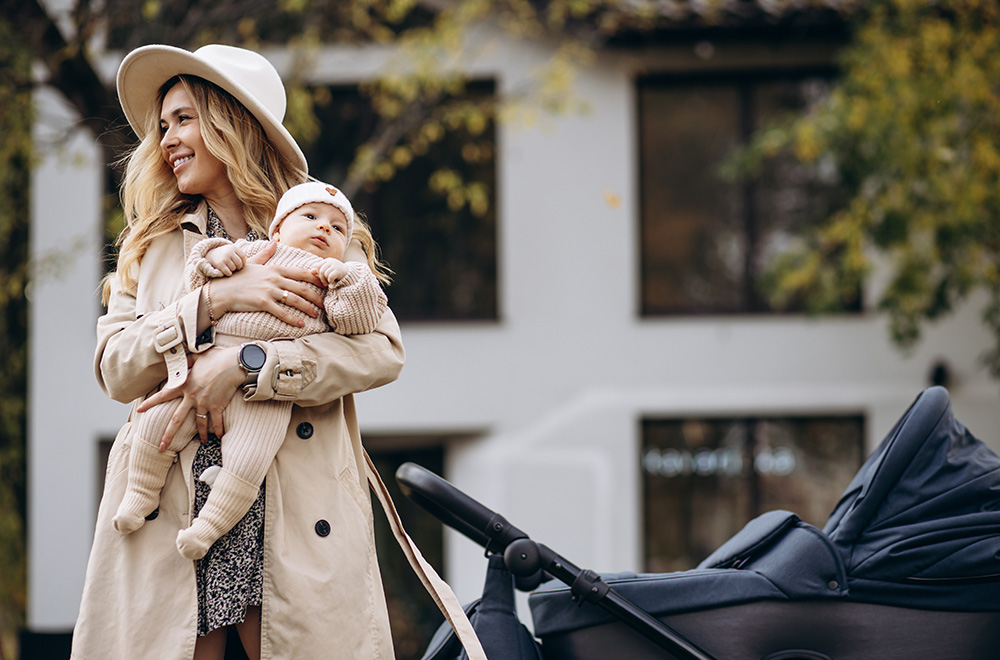 Una mujer sostiene a un bebé en brazos mientras camina junto a un cochecito frente a un edificio.